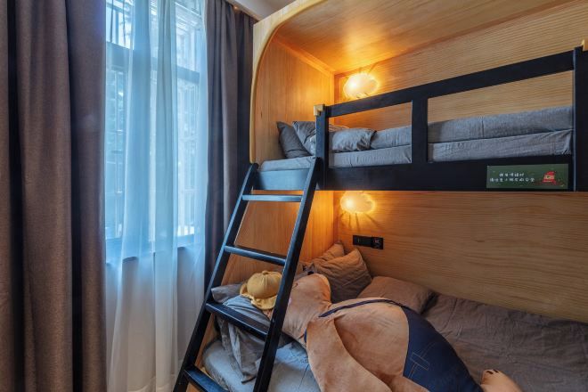 Una cama litera con escalera, una lámpara fijada a la pared y una ventana con cortinas.