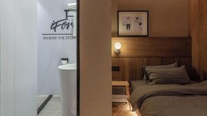 Ένα σύγχρονο δωμάτιο ξενοδοχείου με ξύλινο προσκέφαλο, ένα κρεβάτι με γκρι κλινοσκεπάσματα, ένα κομοδίνο με ένα φωτιστικό και μια κορνιζαρισμένη εικόνα στον τοίχο.