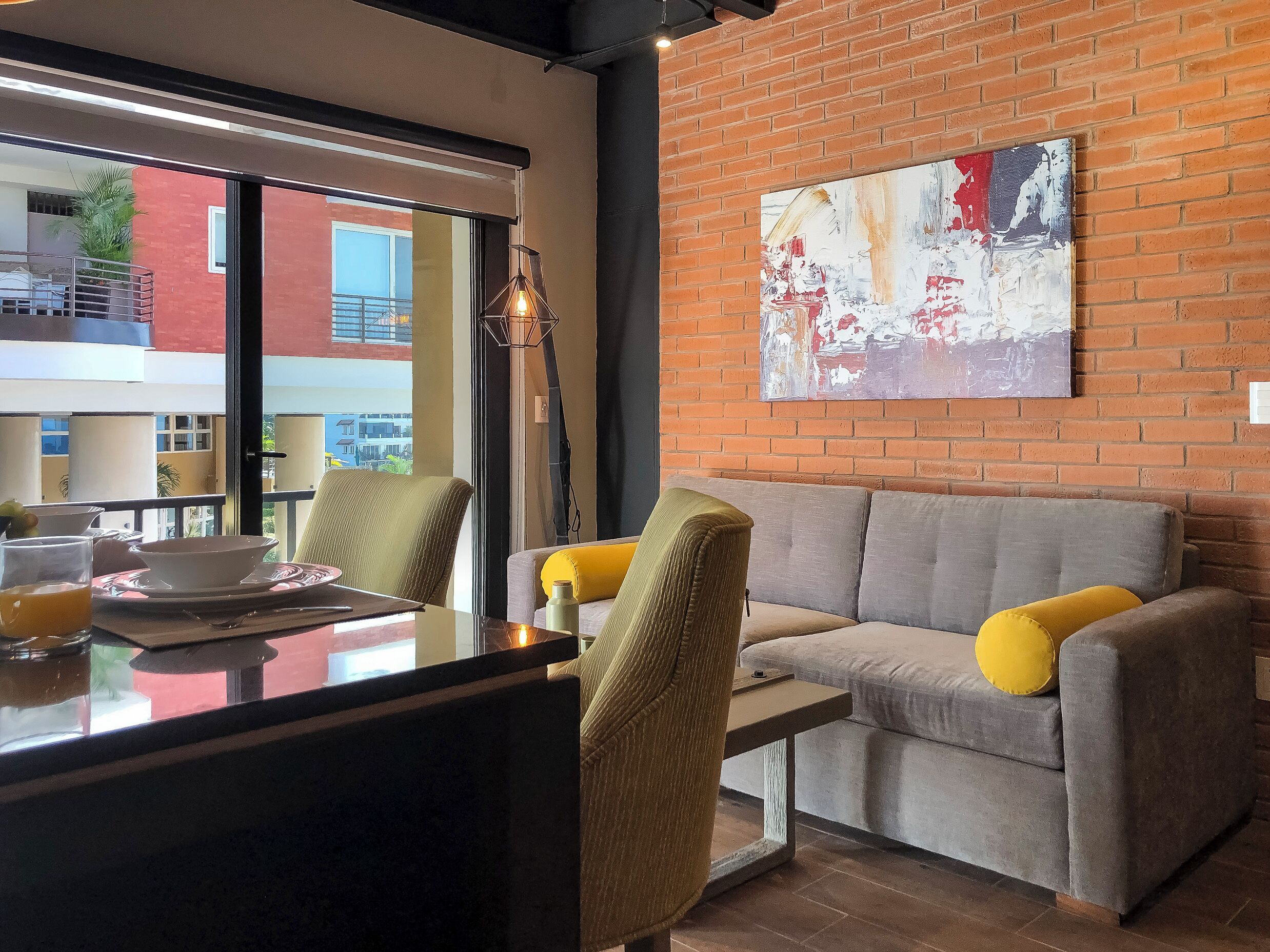 A modern living area with a sofa, chairs, a dining table, and a large abstract painting on a brick wall.
