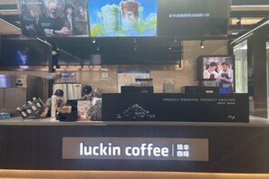 A coffee shop interior with a counter displaying 'luckin coffee' signage, a coffee machine, and a television screen above the counter. - Zhengzhou Xinyang Lingrui Building Hotel (Provincial People's Hospital Metro Station) (Zhengzhou)