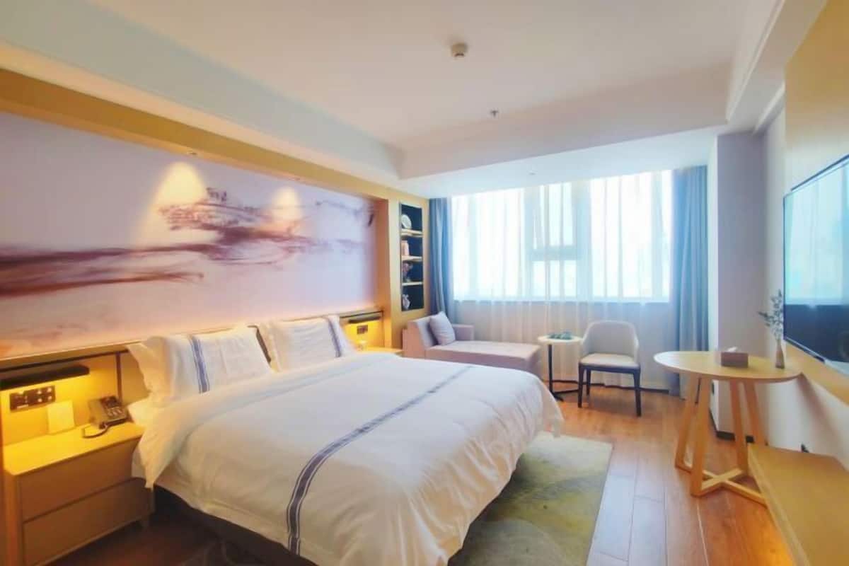 A hotel room with a large bed, bedside tables, a desk, a chair, a TV, and a large window with curtains.
