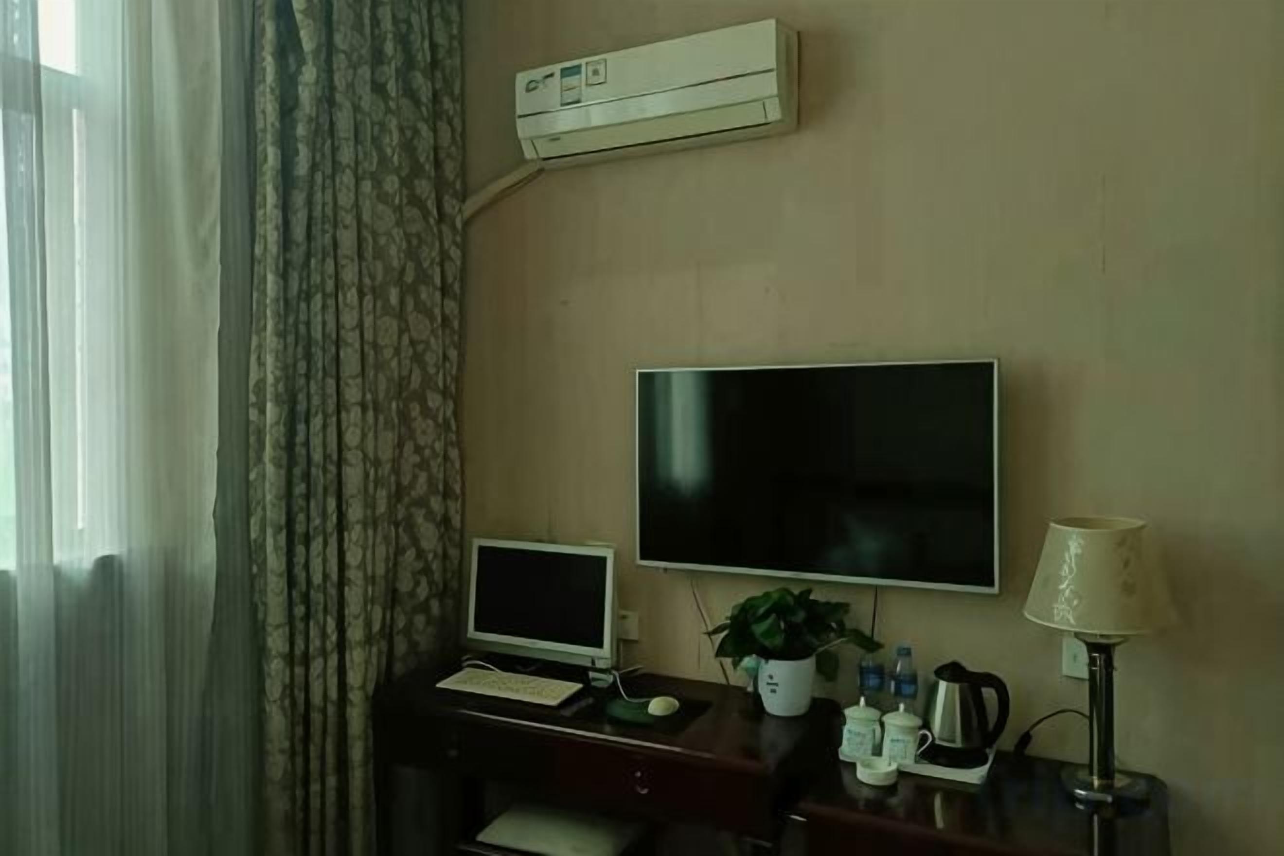 A hotel room with a desk, a flat-screen TV, a laptop, a potted plant, a lamp, and an air conditioning unit.