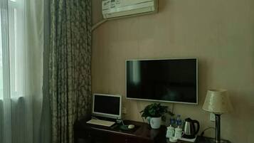 A hotel room with a desk, a flat-screen TV, a laptop, a potted plant, a lamp, and an air conditioning unit.