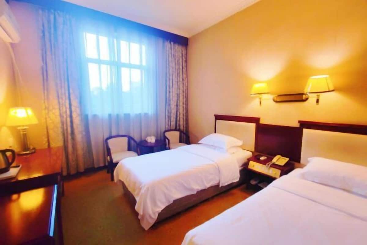 A hotel room with two beds, a desk with a lamp, a chair, a nightstand with a phone, and a window with curtains.