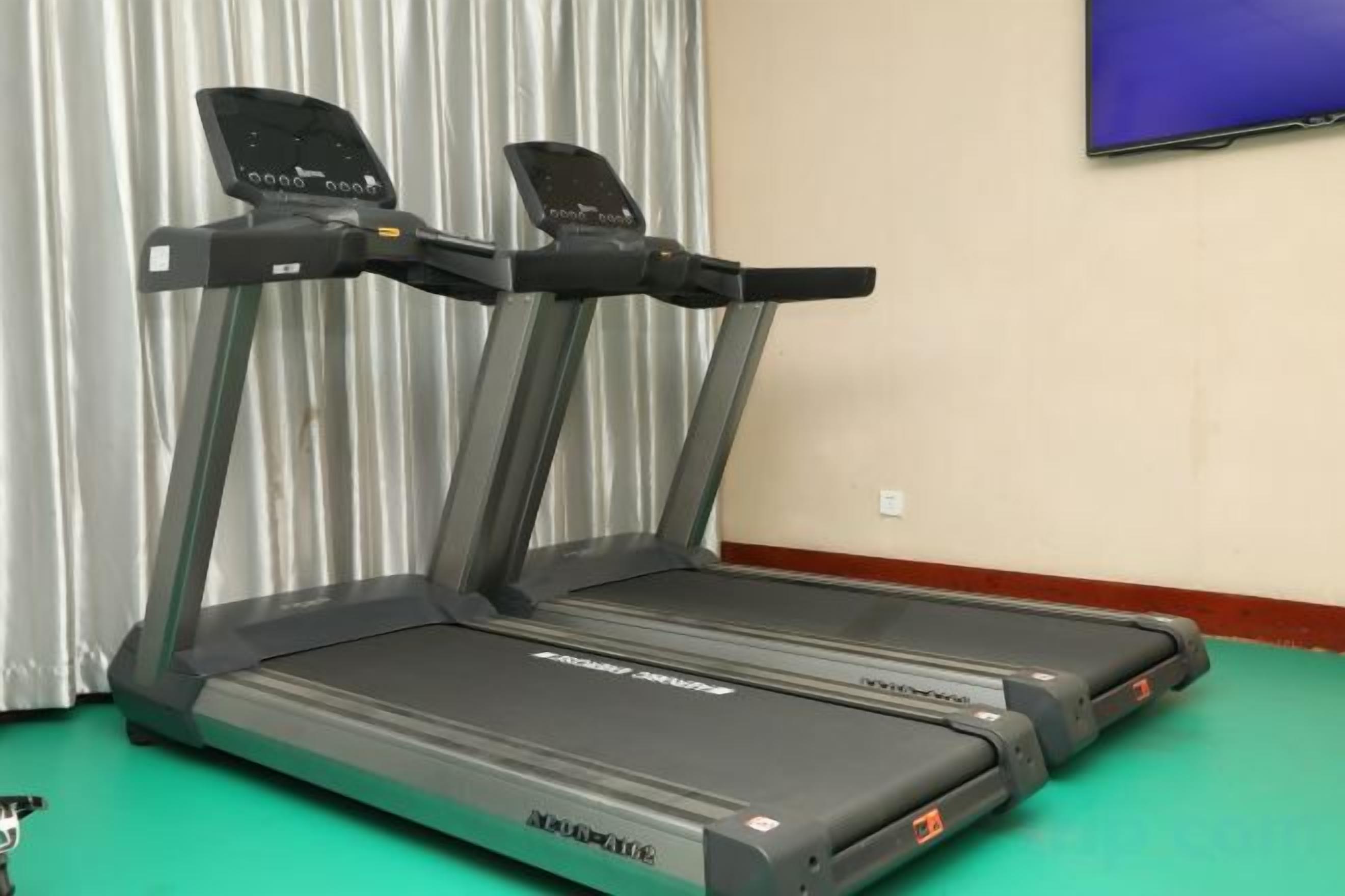 Una caminadora en una habitación con piso verde, un televisor en la pared y una ventana con cortinas.