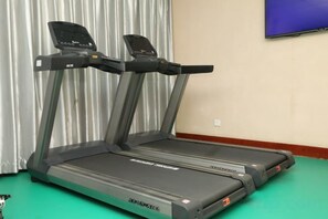 A treadmill in a room with a green floor, a television on the wall, and a window with curtains. - Zhengzhou Xinyang Lingrui Building Hotel (Provincial People's Hospital Metro Station) (Zhengzhou)