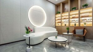 Un espace d’accueil moderne avec un canapé blanc aux lignes courbes, un miroir rond et une bibliothèque agrémentée de plantes. On y trouve également deux petites tables rondes et un fauteuil en cuir marron.