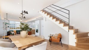Ein modernes Wohnzimmer mit einem Holzesstisch, weißen Stühlen, einer Couch und einer Treppe mit Holzstufen und einem Metallgeländer.