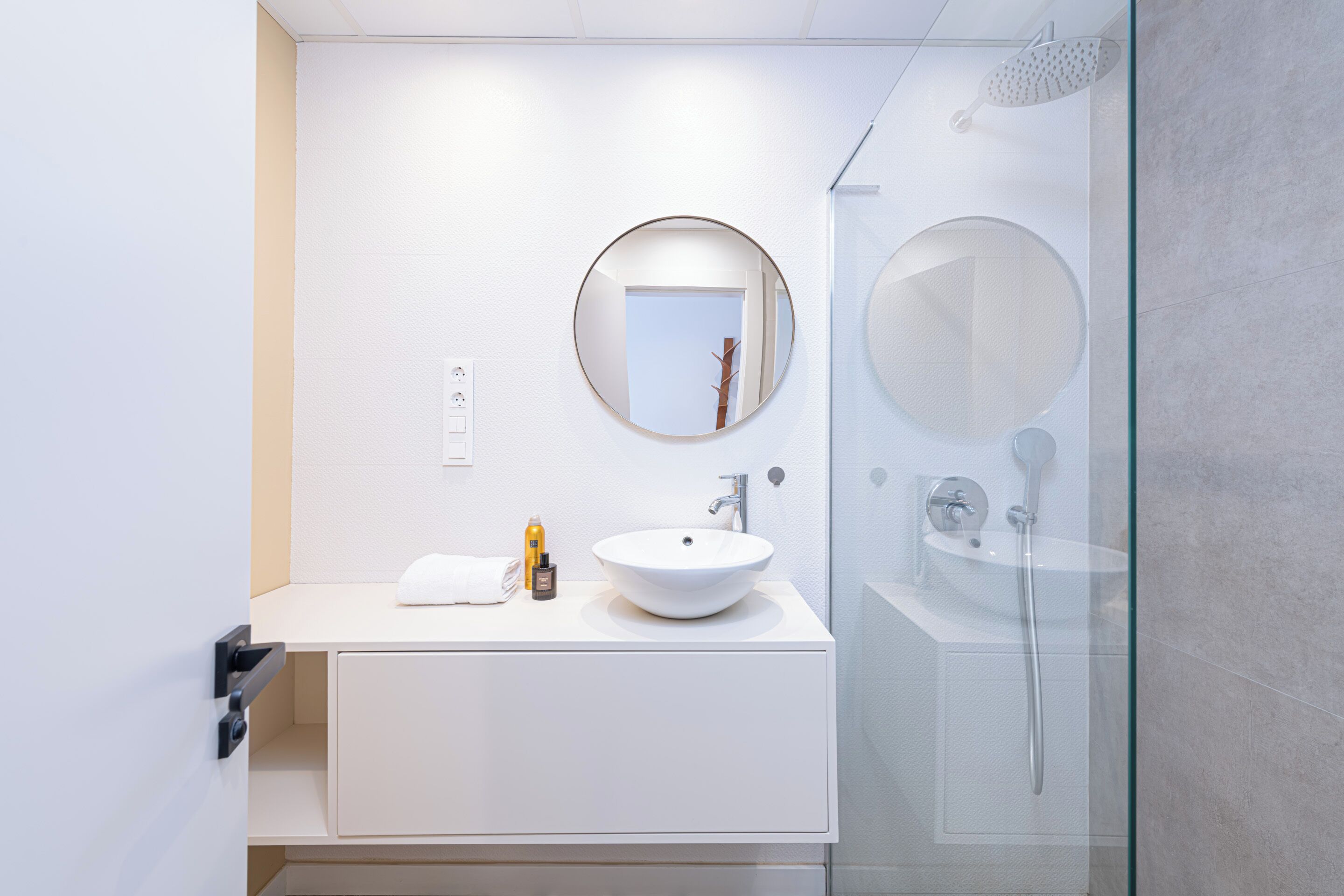 A modern bathroom with a white sink, round mirror, and glass shower enclosure.