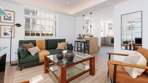 Ein geräumiger Wohnbereich mit einem grünen Sofa, einem Glastisch und einem braunen Sessel. Im Hintergrund ist eine Küchenzeile mit einem Barhocker und einem Esstisch erkennbar.
