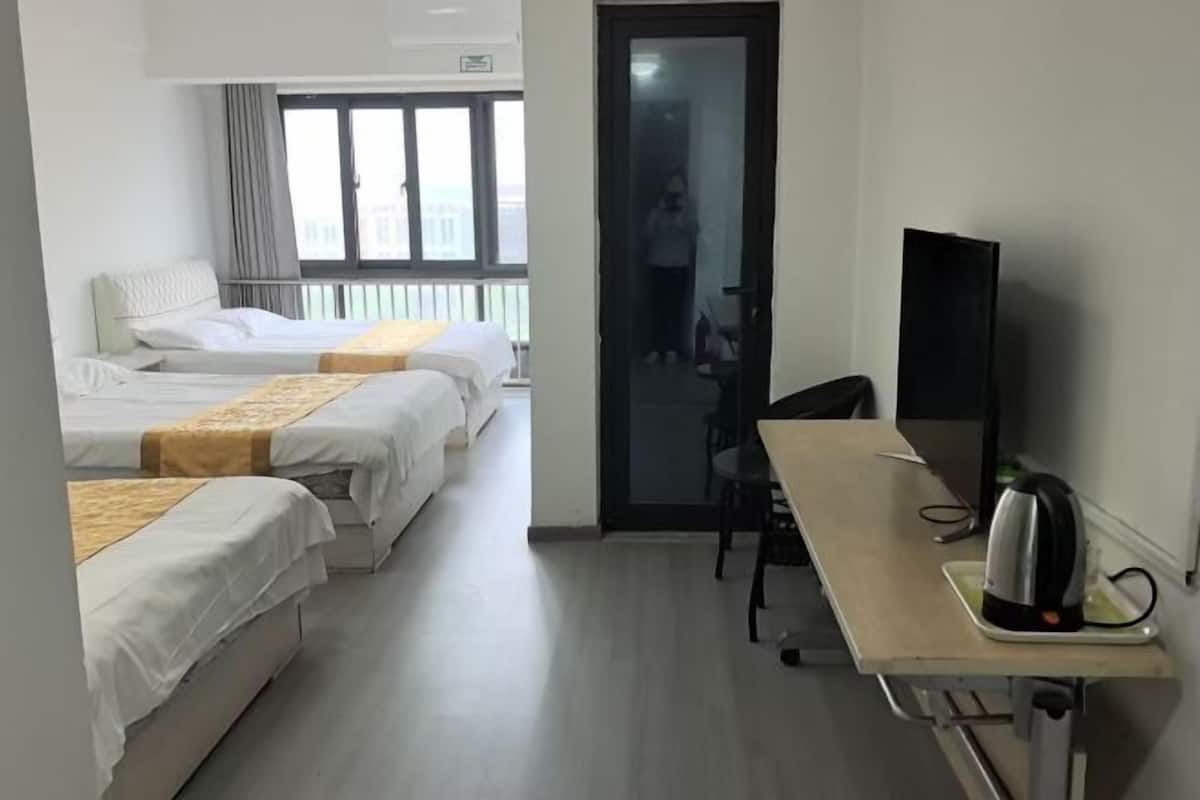A hotel room with three beds, a desk with a kettle, a chair, and a TV.