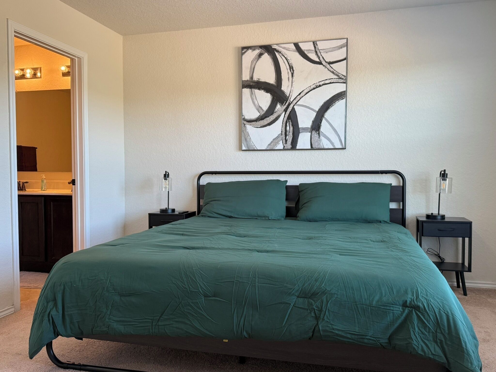 A bedroom with a bed, two bedside tables, a wall art piece, and a bathroom visible through an open door.
