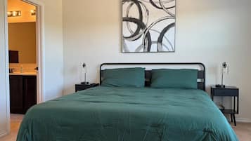 A bedroom with a bed, two bedside tables, a wall art piece, and a bathroom visible through an open door.