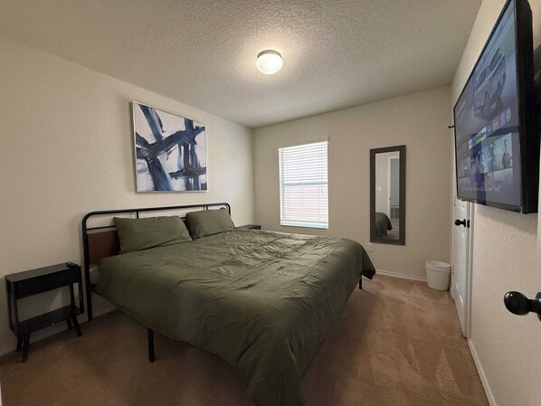 A bedroom with a bed, a nightstand, a wall-mounted TV, a window with blinds, and a ceiling light.