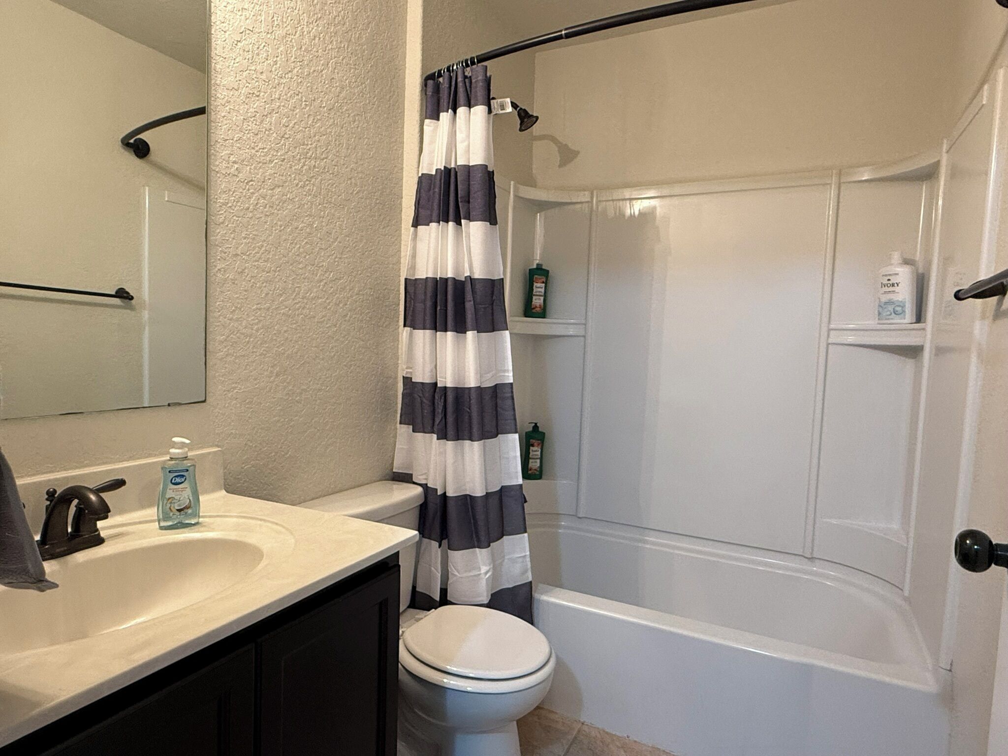 Een badkamer met een witte wastafel, een toilet, een douche met een zwart-wit gestreept gordijn, en toiletartikelen op de muur.