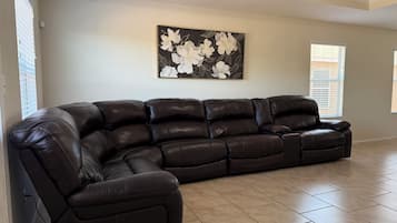 A spacious living room with a large L-shaped sectional sofa, a wall-mounted floral artwork, and large windows with blinds.