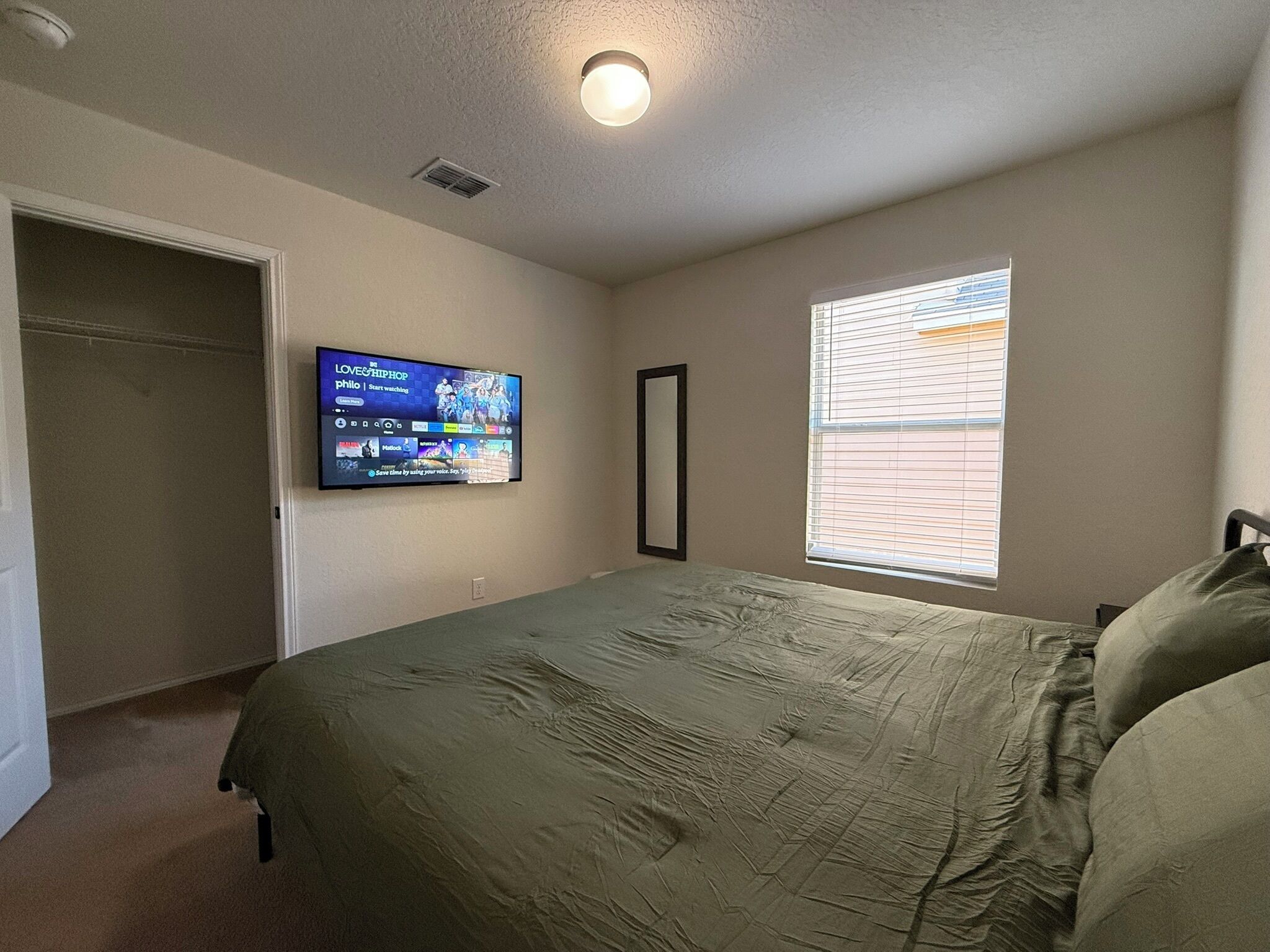 A bedroom with a bed, a TV mounted on the wall, a window with blinds, and a door.