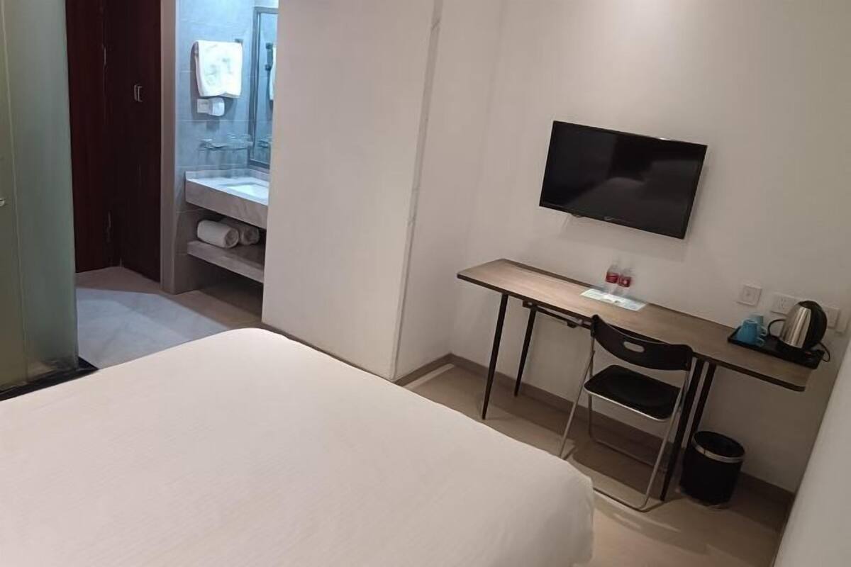 A hotel room with a bed, a desk with a chair, a television, and a small trash can.
