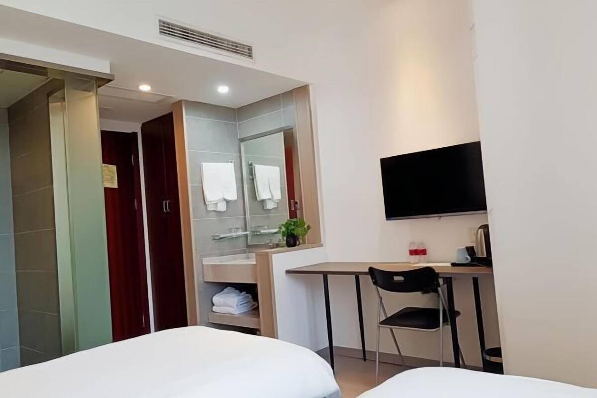 A hotel room with two beds, a desk, a chair, a TV, and a bathroom with a mirror and towel rack.