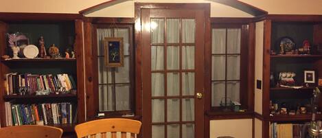 A cozy room with a wooden door, bookshelves, a dining table with chairs, and a ceiling fan.