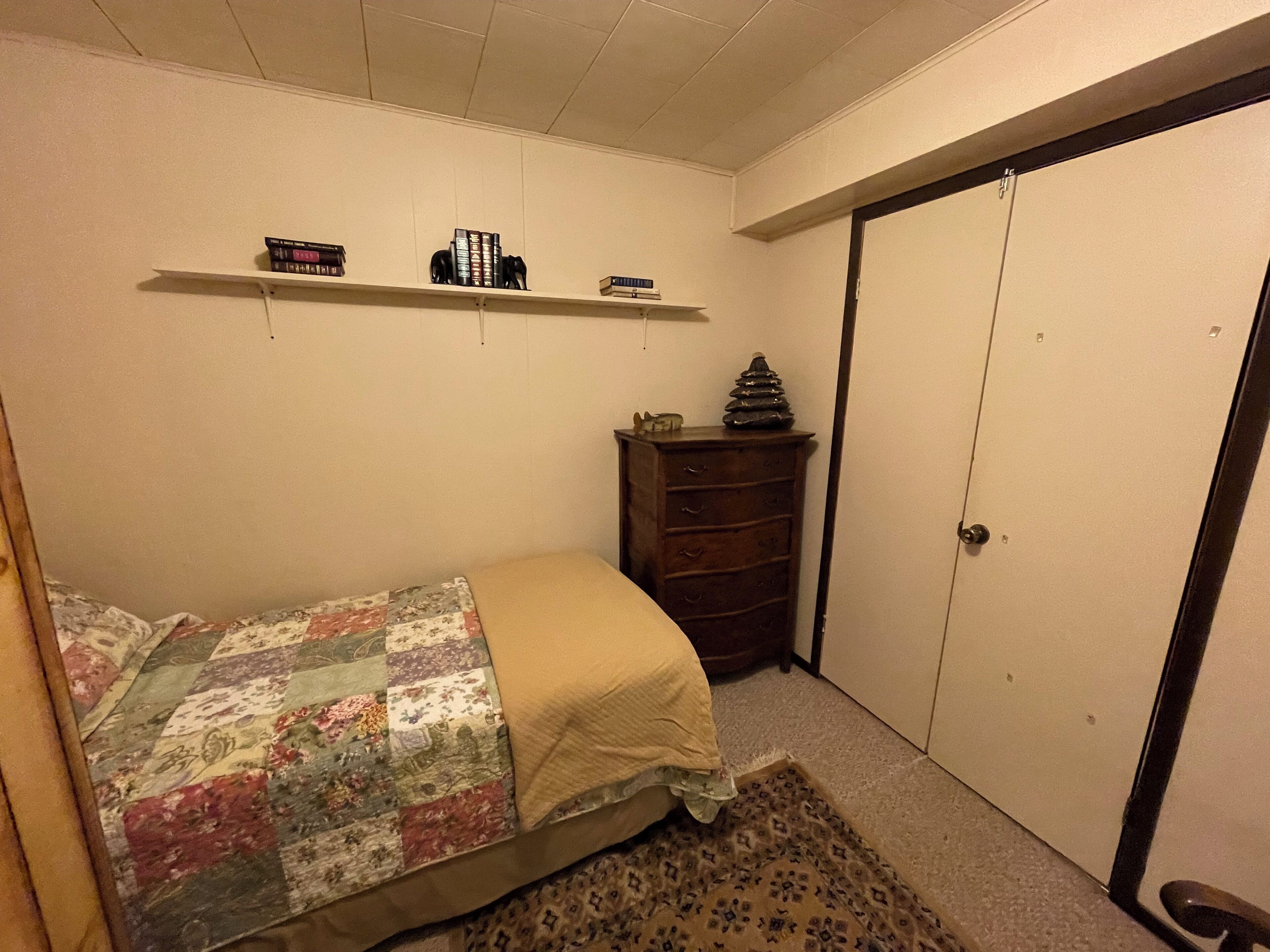 A bedroom with a bed, a dresser, a wardrobe, and shelves with books and a decorative item.