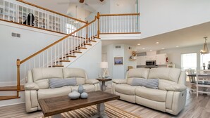 A spacious living room with a wooden staircase, two beige sofas, a coffee table, and a kitchen in the background. - Waterfront ~ Ohana  ~ Sleeps 10 (Dauphin Island)