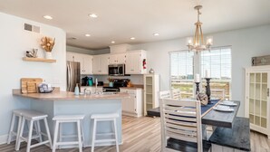 A modern kitchen with white cabinets, a central island with stools, and a dining area with a table and chairs. - Waterfront ~ Ohana  ~ Sleeps 10 (Dauphin Island)