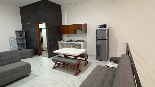 Un salon moderne avec un canapé gris, un banc en bois et une table à manger. Il y a également un coin cuisine équipé d’un réfrigérateur, d’un four à micro-ondes et de placards.