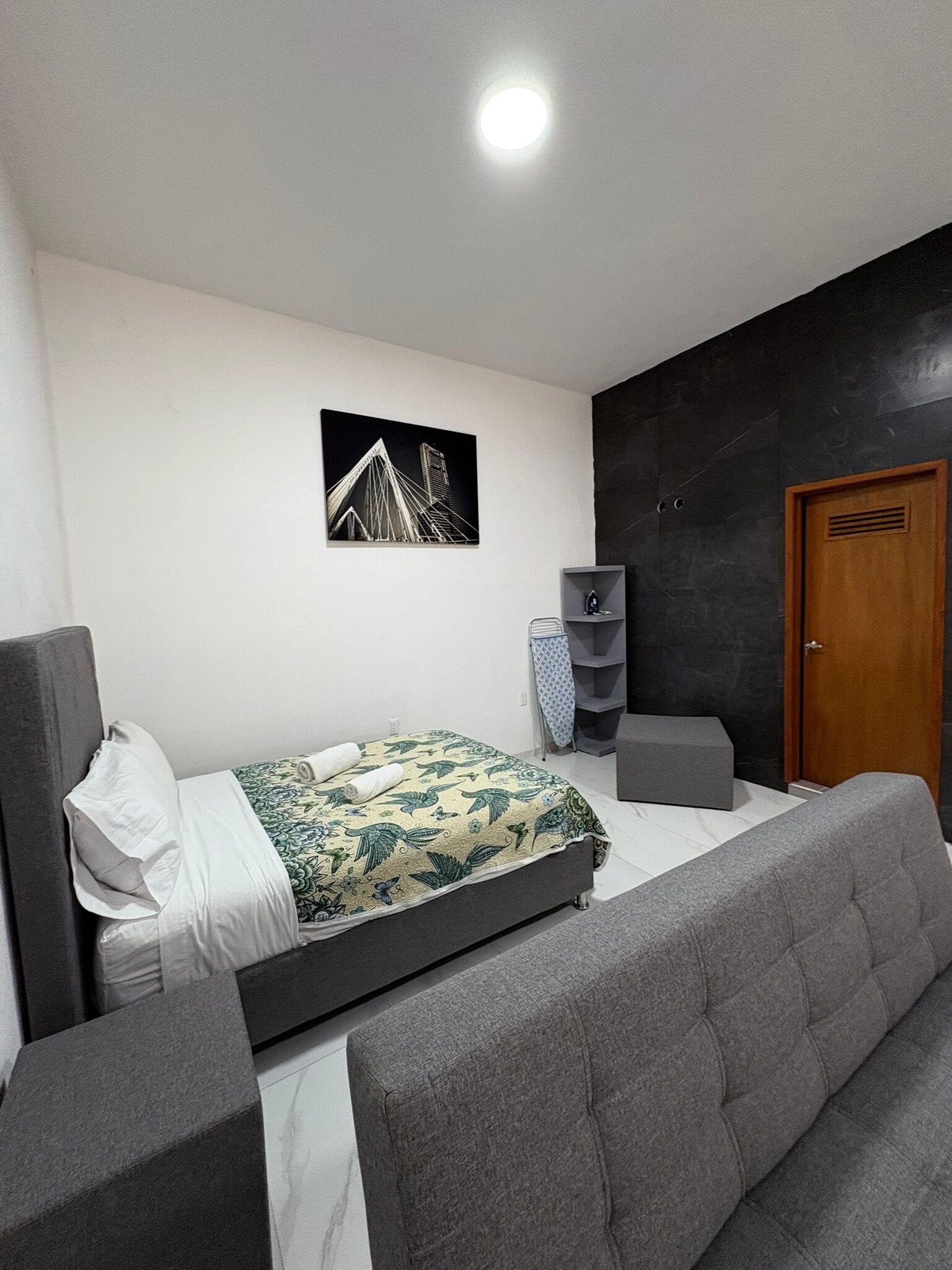 Een moderne slaapkamer met een bed, een grijze bank, een houten deur en een zwarte muur met een ingelijst kunstwerk.