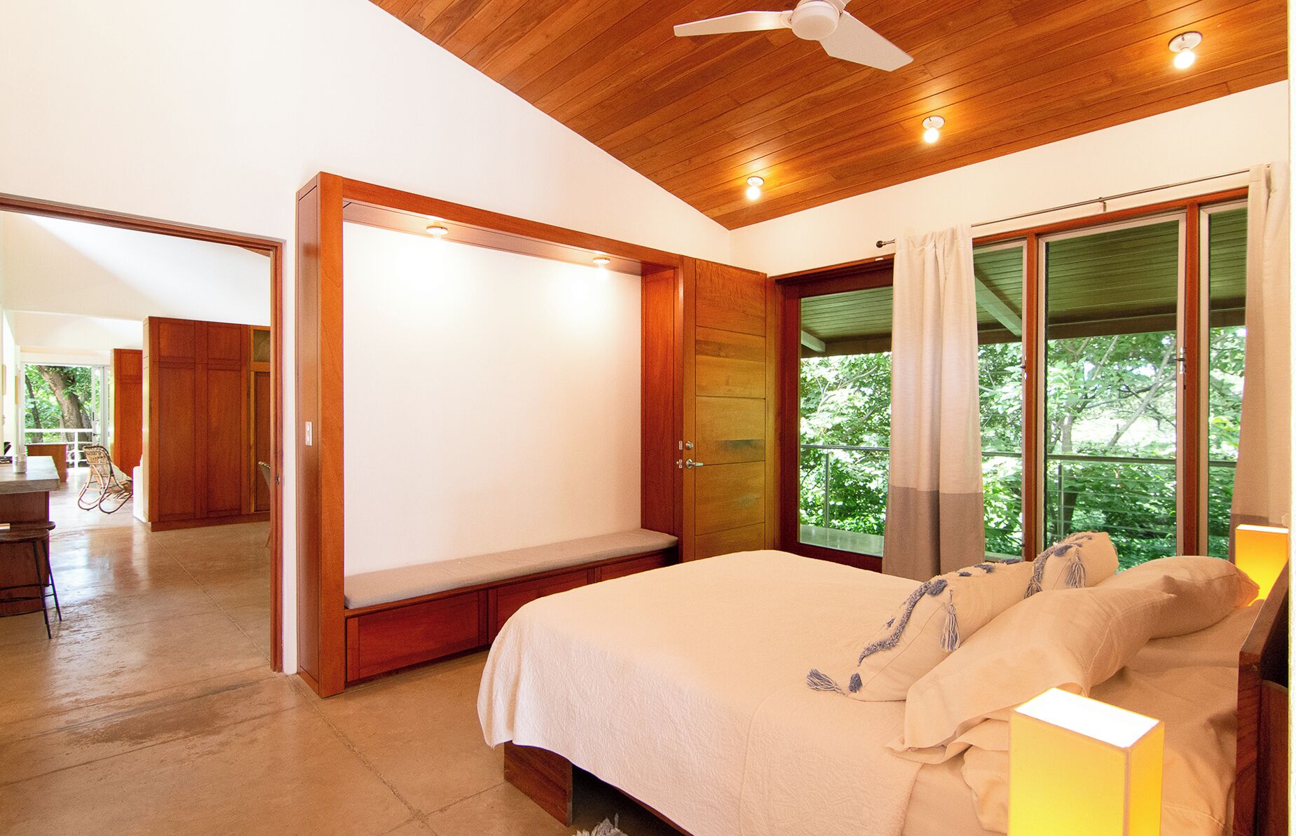 A bedroom with a bed, a ceiling fan, a window with curtains, and a view of greenery outside.