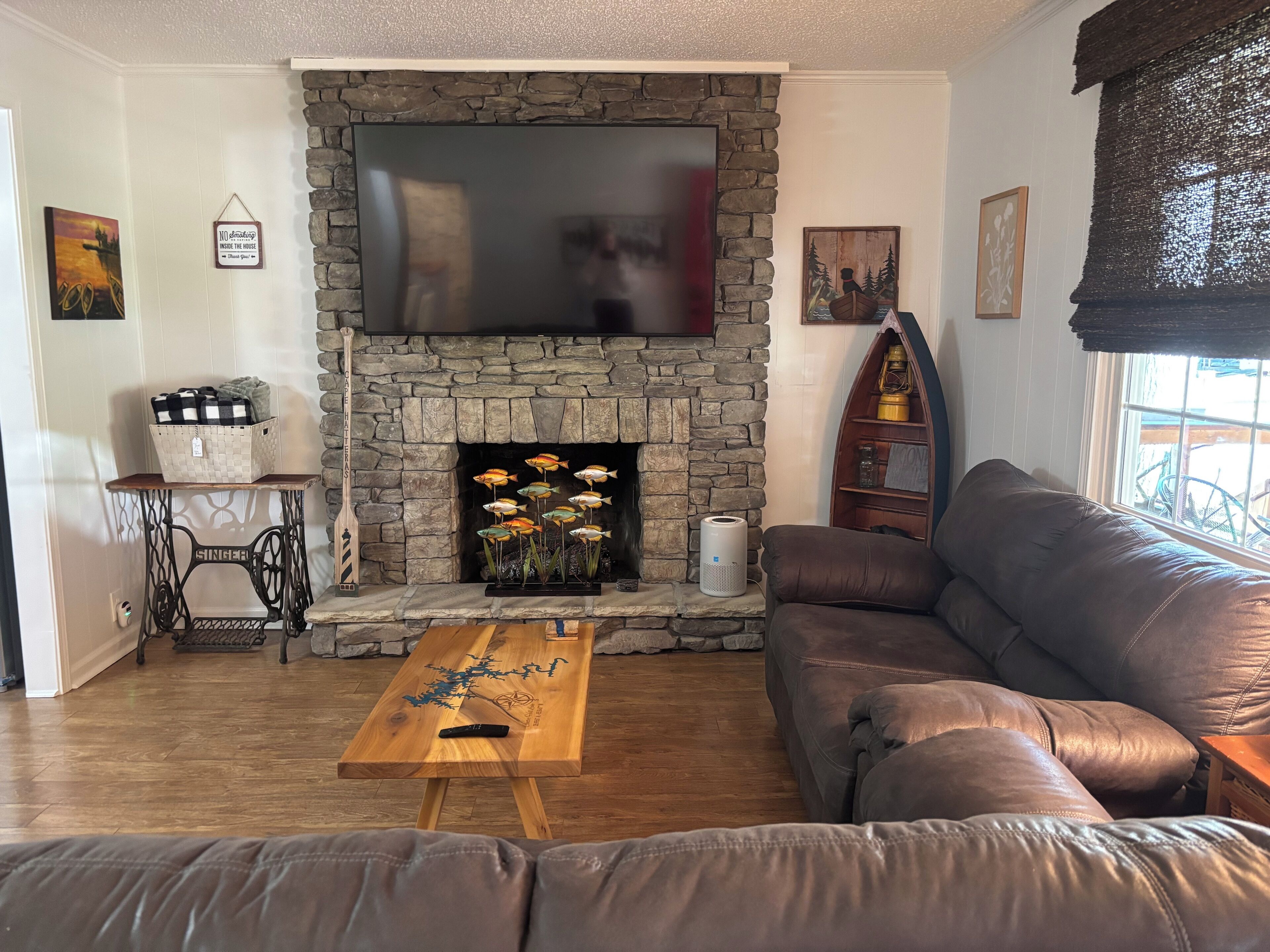 A living room with a stone fireplace, a television mounted above it, a brown leather sofa, a wooden coffee table, and a small side table with a basket.
