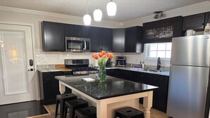 Una cocina moderna con armarios oscuros, un refrigerador de acero inoxidable, un microondas y una estufa a gas. Hay una encimera negra con un jarrón de tulipanes y dos taburetes de bar.