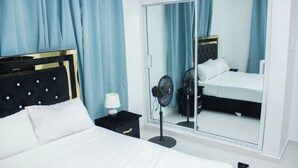 A hotel room with a bed, a bedside table, a lamp, a fan, and a large mirror.