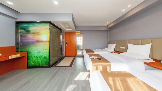 Ein Hotelzimmer mit einem großen digitalen Bildschirm, der einen Sonnenuntergang zeigt, einem Bett mit weißer Bettwäsche, einem Holztisch und einem Empfangsbereich mit Glastür.