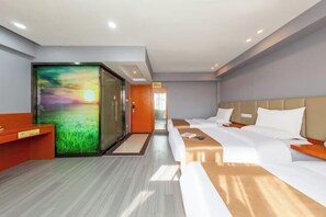 Habitación de hotel con una gran pantalla digital mostrando un atardecer, una cama con ropa blanca, un escritorio de madera y una zona de recepción con puerta de vidrio.