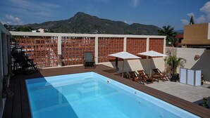 A swimming pool with wooden deck and lounge chairs, surrounded by a wooden fence and palm trees, with mountains in the background.