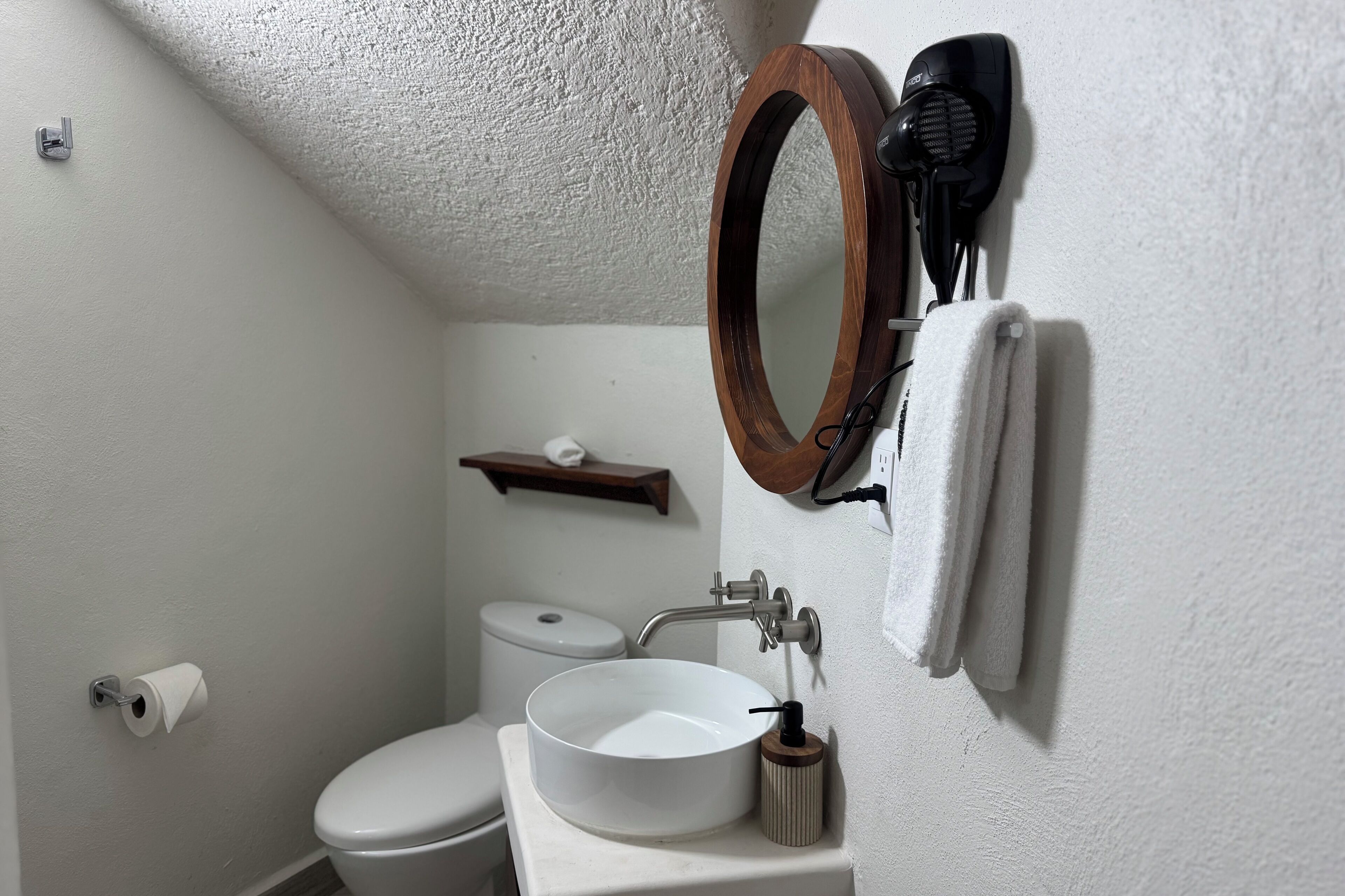 Un baño con un lavamanos blanco, un espejo redondo, una toalla colgada en la pared, un inodoro y un estante con un rollo de papel higiénico.
