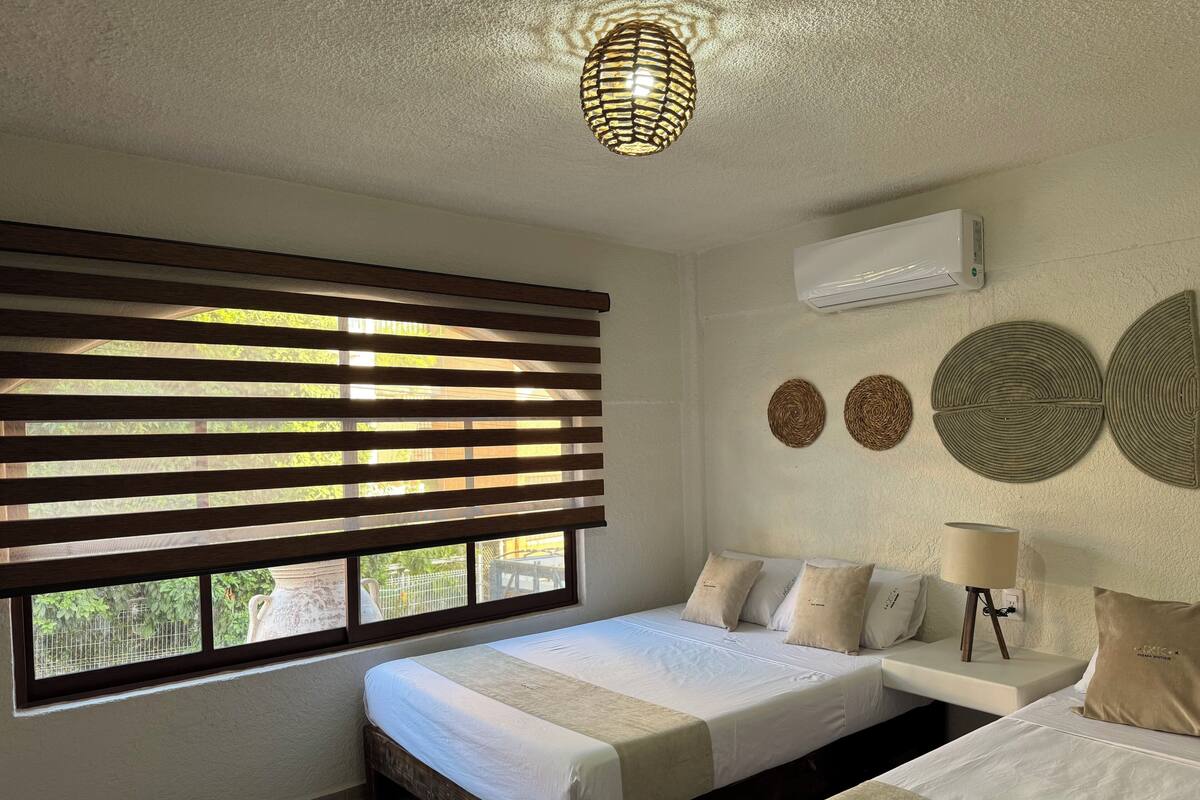 A bedroom with two beds, a window with blinds, a wall-mounted air conditioner, and decorative wall art.