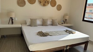 A neatly made bed with a white mattress and beige bedding, a bedside table with a lamp, and a wall decorated with woven wall art.