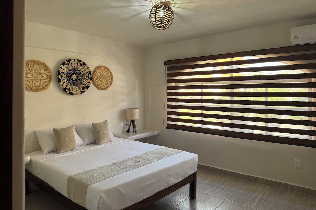 A bedroom with a large bed, a bedside table with a lamp, and a window with blinds.