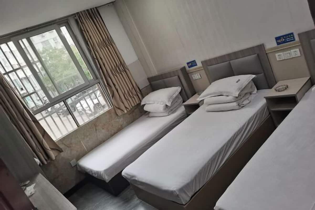 Une chambre d’hôtel avec deux lits simples, une fenêtre avec des rideaux, une table de chevet avec une lampe et une prise de courant fixée au mur.