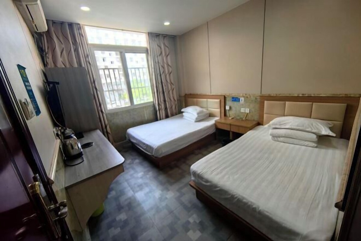 Ein Hotelzimmer mit zwei Einzelbetten, einem kleinen Schreibtisch mit Wasserkocher, einem Fenster mit Vorhängen und einer Wand-Klimaanlage.