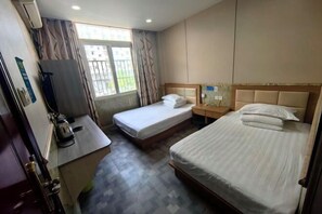 Ein Hotelzimmer mit zwei Einzelbetten, einem kleinen Schreibtisch mit Wasserkocher, einem Fenster mit Vorhängen und einer Wand-Klimaanlage.
