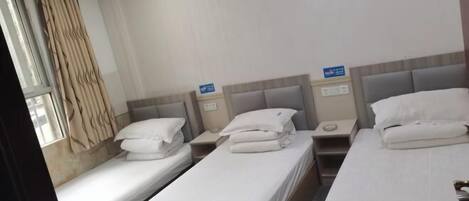A hotel room with three single beds, each with white linens and pillows, a bedside table, and a nightstand.