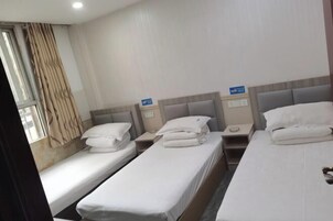 A hotel room with three single beds, each with white linens and pillows, a bedside table, and a nightstand.