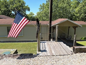 A wooden deck leading to a covered porch with a metal gate, an American flag, and a house with a red roof.
