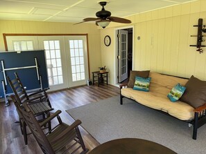 A room with a sofa, wooden chairs, a ceiling fan, a small table with a vase, and a door leading to another room.