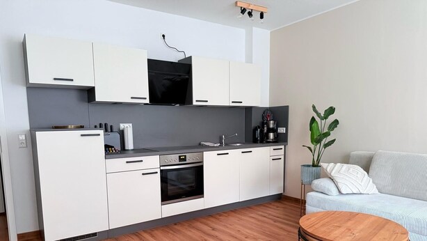 Una cocina moderna con armarios blancos, horno empotrado y cafetera. Hay una mesa redonda de madera con patas metálicas y un sofá a rayas en la sala de estar.