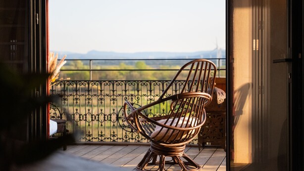 A wicker chair on a balcony with a view of a fence and greenery.