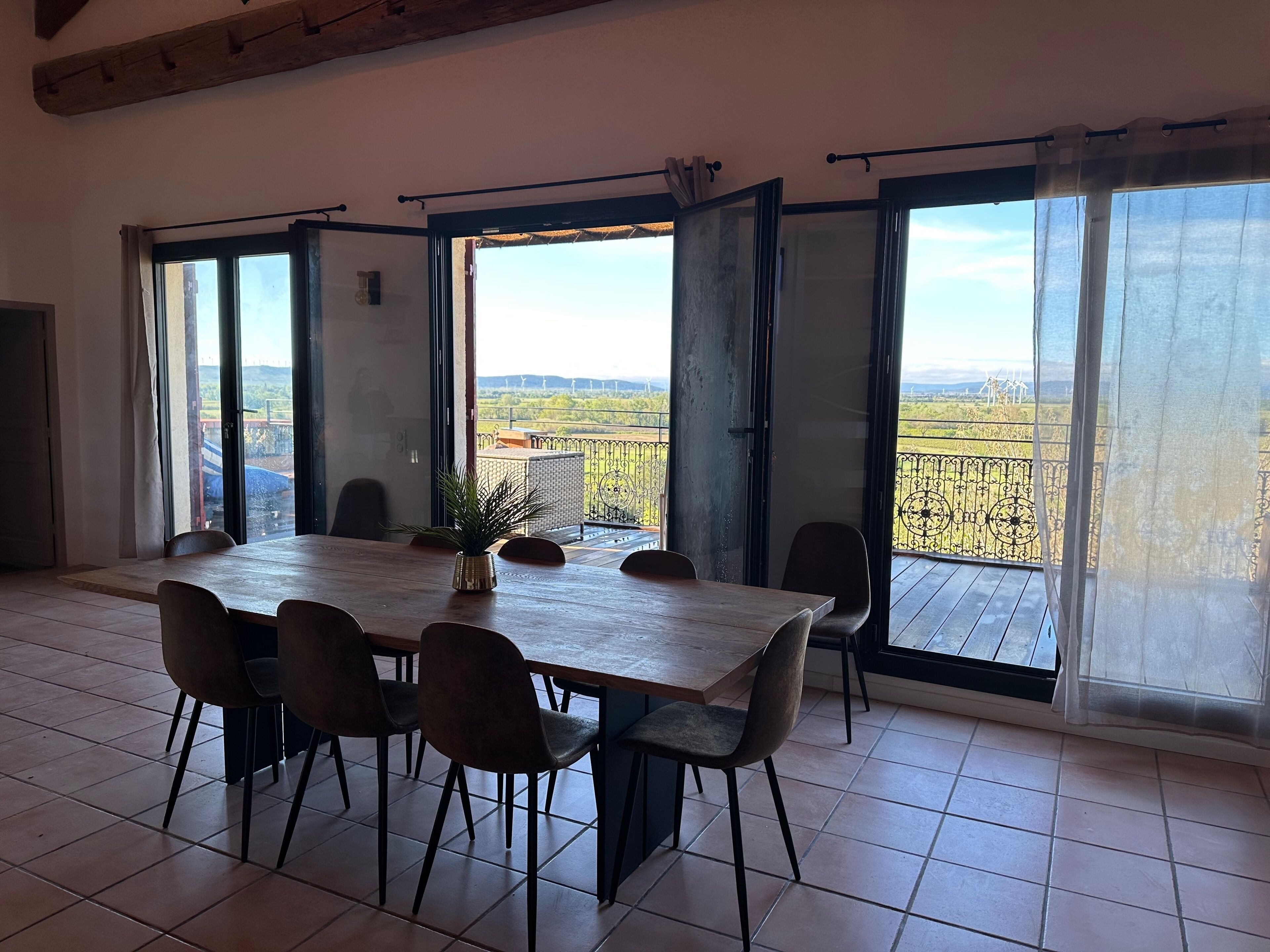Ein Essbereich mit großem Tisch und Stühlen, großen Fenstern mit Blick auf Windkraftanlagen und einem Balkon mit Metallgeländer.